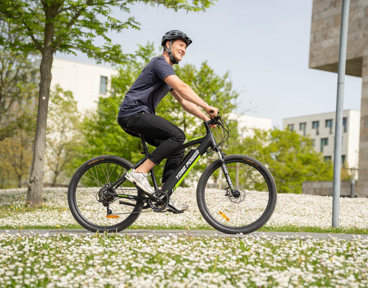 Man riding a YOSE POWER electric mountain bike through a sunny, flower-filled park, wearing a helmet and smiling.
