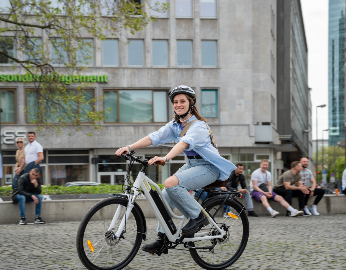 Woman riding a YOSE POWER electric city bike on a street in an urban area, smiling and wearing a helmet.