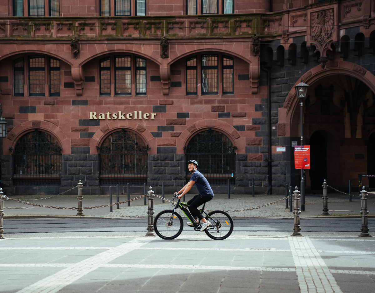 Man riding a YOSE POWER electric mountain bike in front of a building, on a sunny urban street.