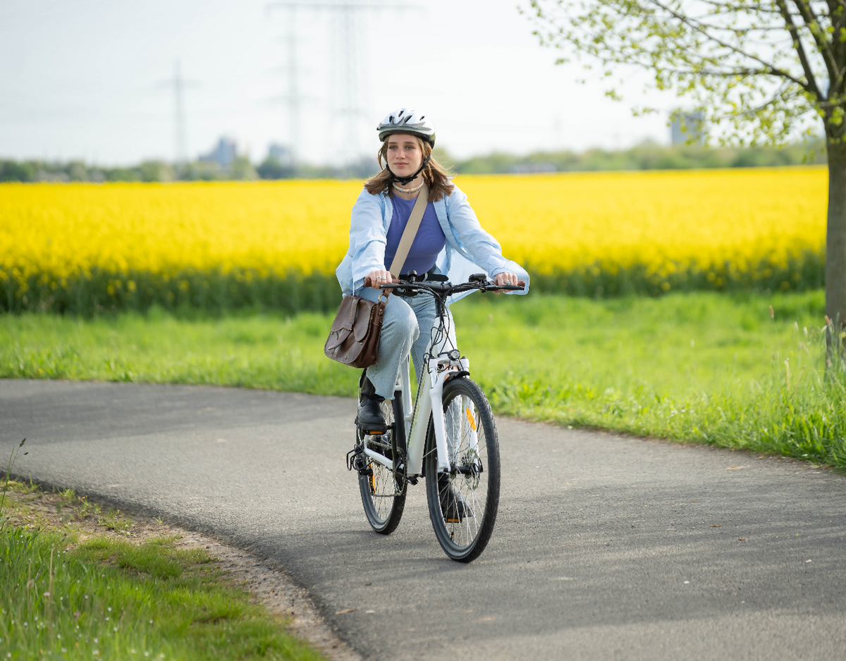 Woman riding a YOSE POWER electric bike on a paved path, with a bright yellow rapeseed field in the background.